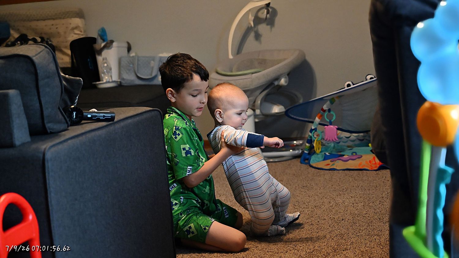 An older child sits on the floor holding a baby near a colorful activity toy indoors.