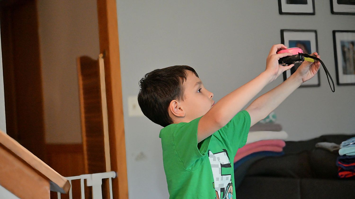 Boy in green shirt holding camera near staircase indoors.