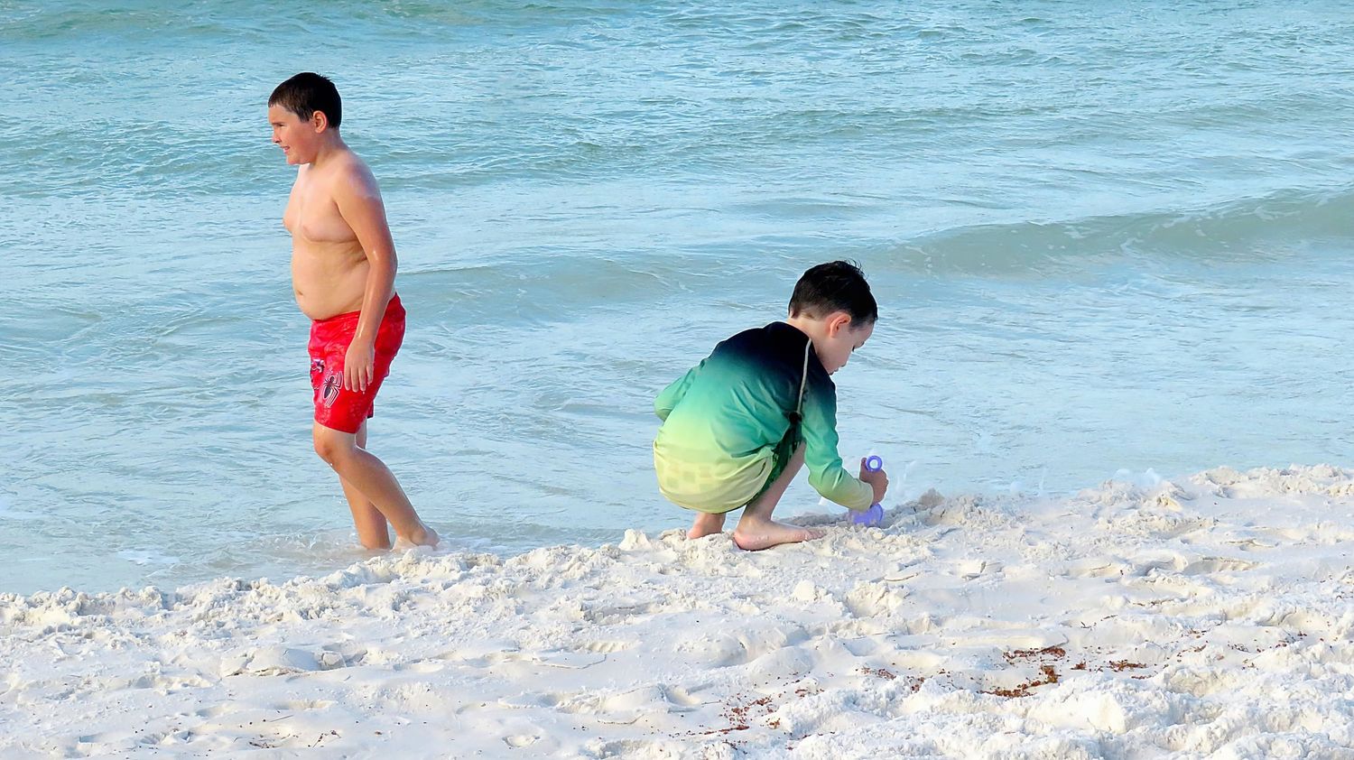 Two boys in swim trunks explore the white sandy shoreline with gentle ocean waves in the background.