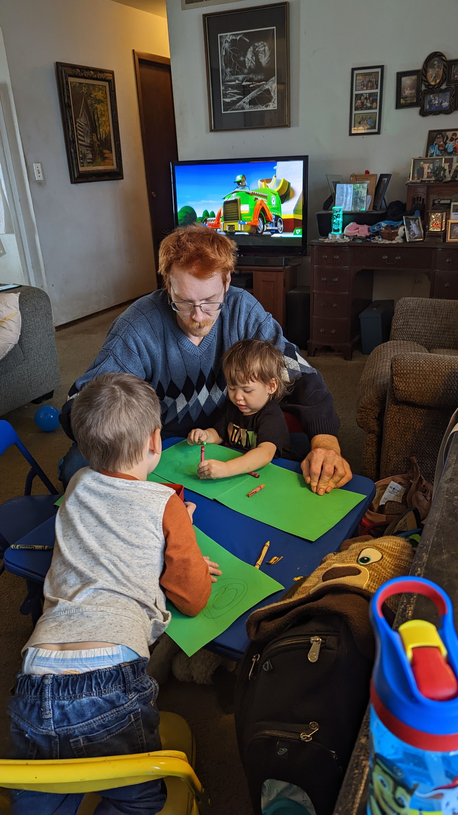 Dad with red hair helps two young boys with art project on green paper at home table with TV in background.