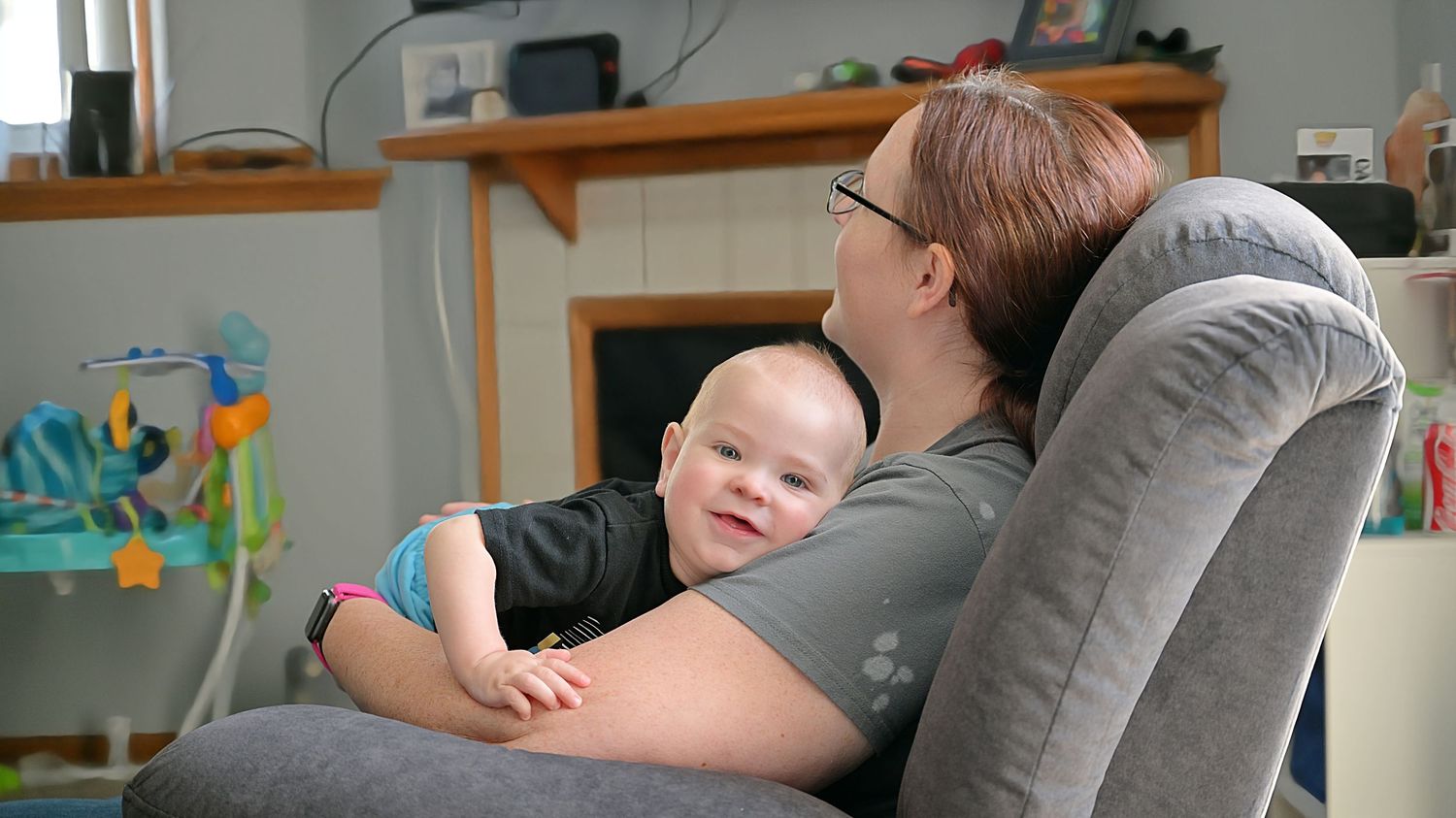 A mother with glasses cuddles her smiling baby in a gray recliner chair in a warm family living room.