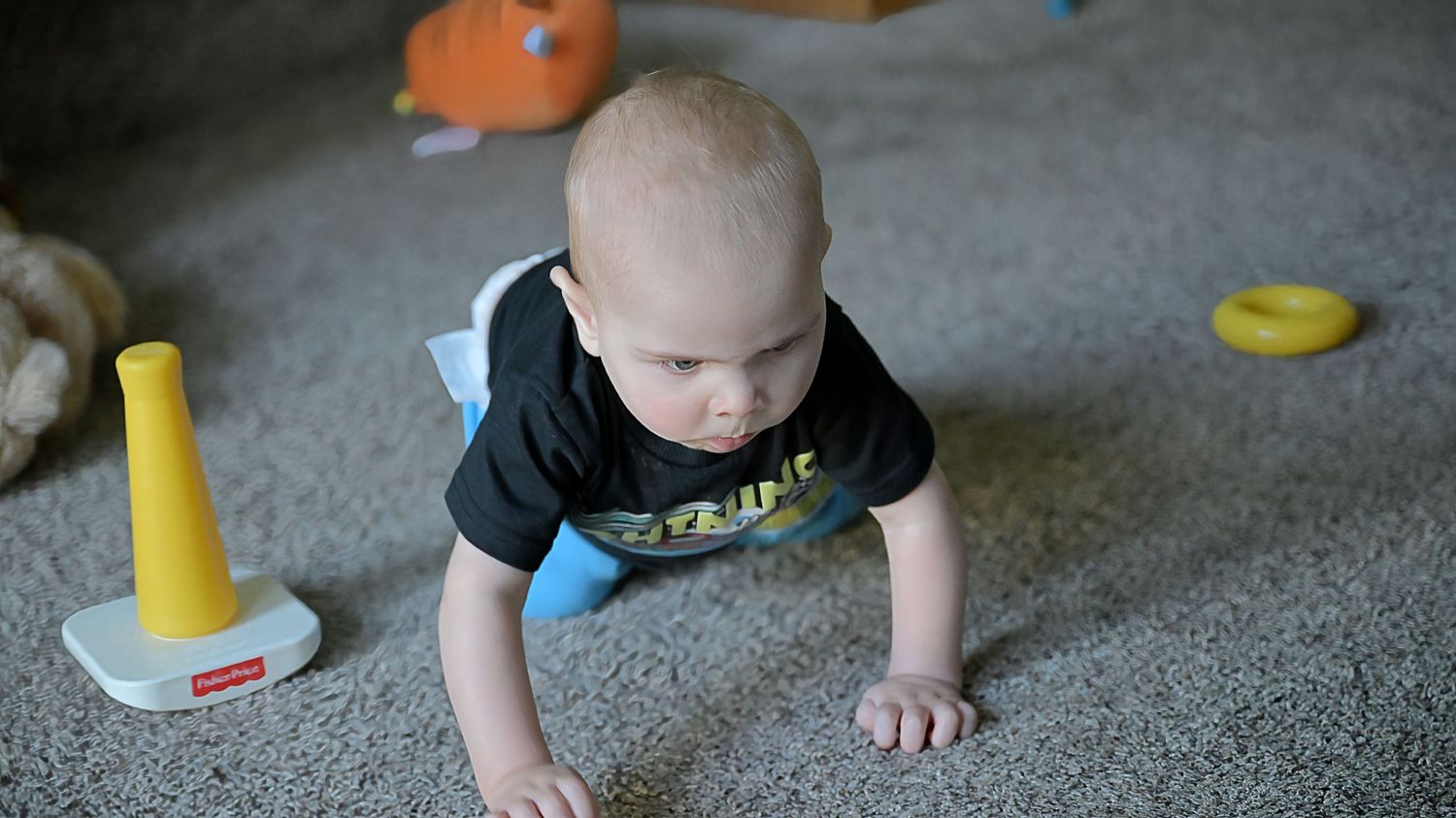 Baby in black shirt crawling on carpet toward yellow and teal toys.