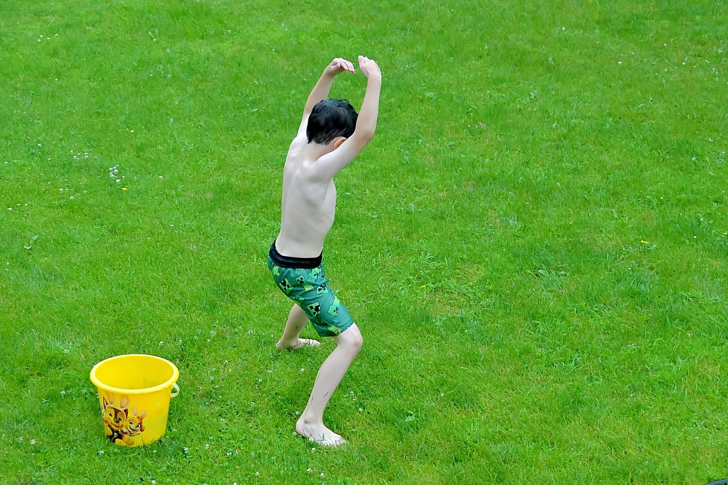 Young boy in swim trunks playing with water next to a yellow bucket.