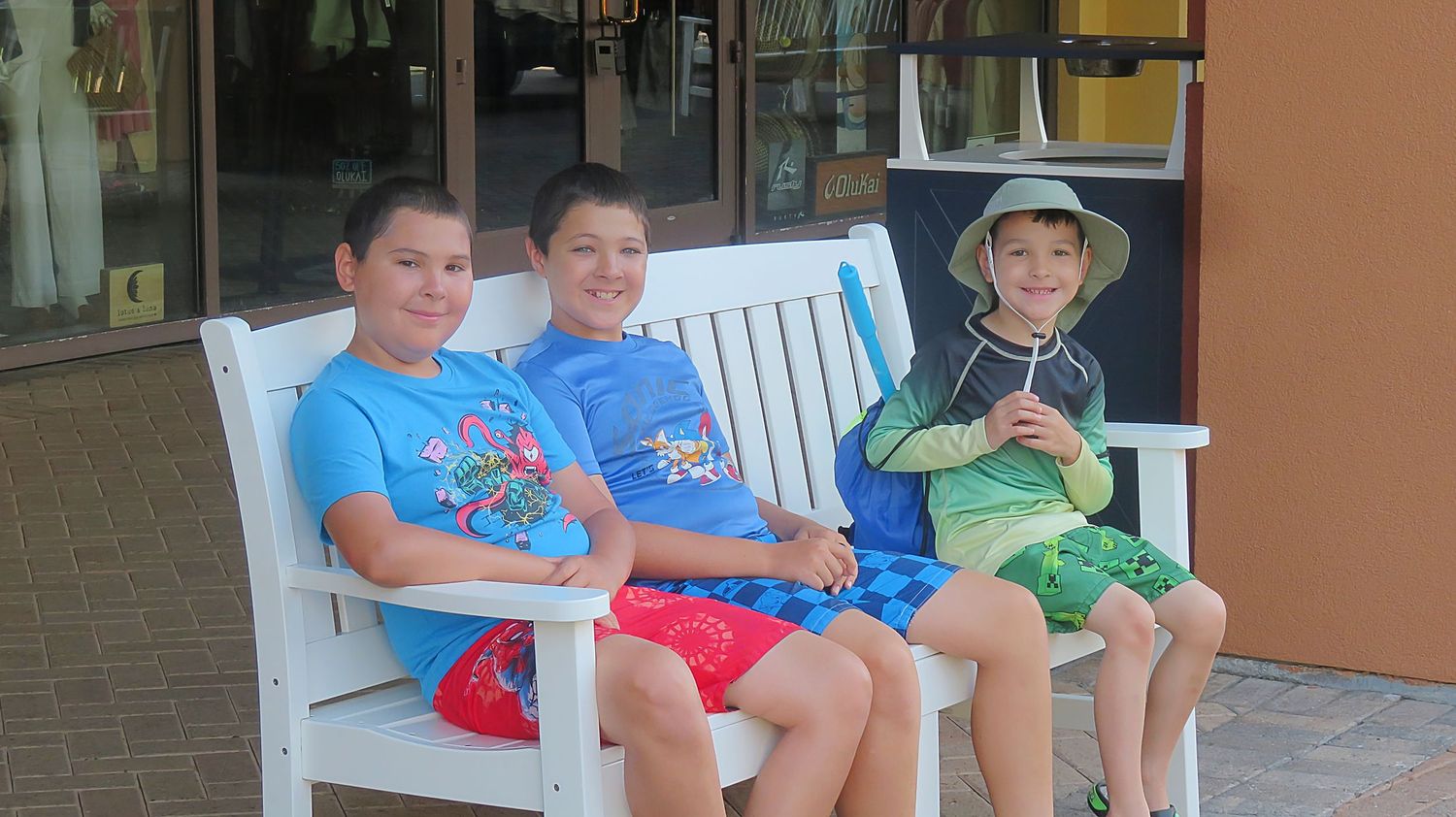 Three boys sitting together on a white wooden bench outside a shop on a sunny day.
