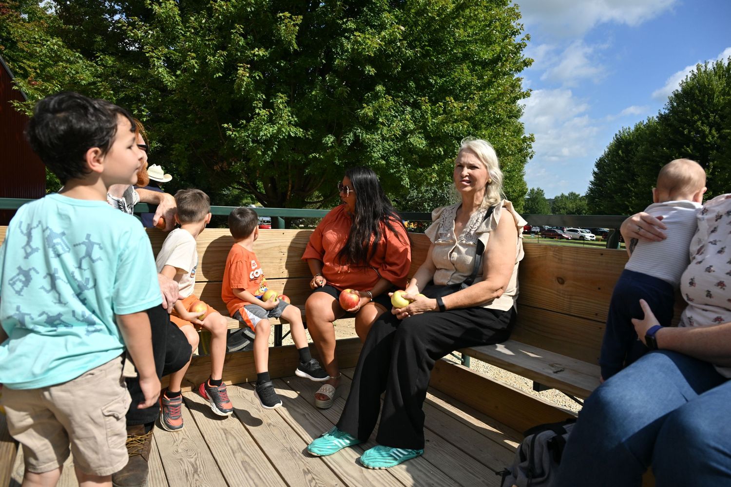 A group of children and adults sit together on wooden benches outdoors surrounded by green trees.