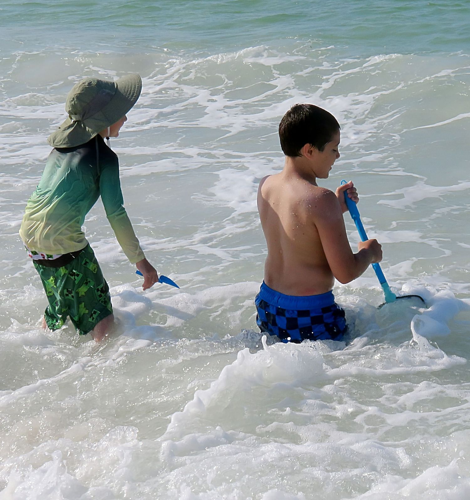 Two boys dig in ocean surf with blue shovels wearing rash guards and swim trunks at beach.