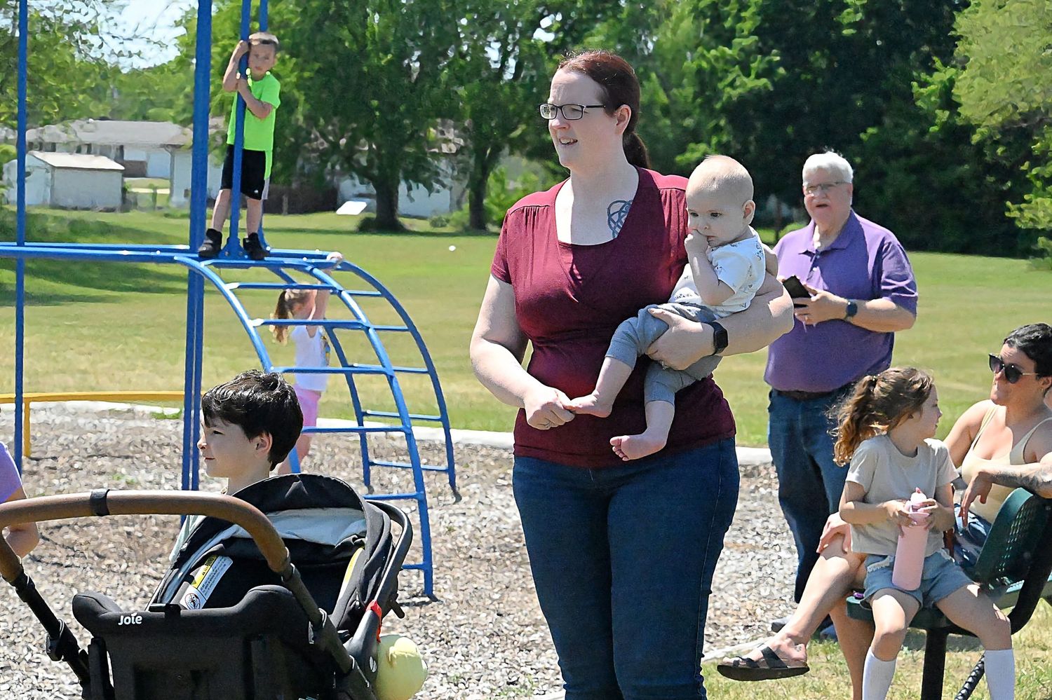 Woman carrying a baby at a sunny playground with children and adults nearby.