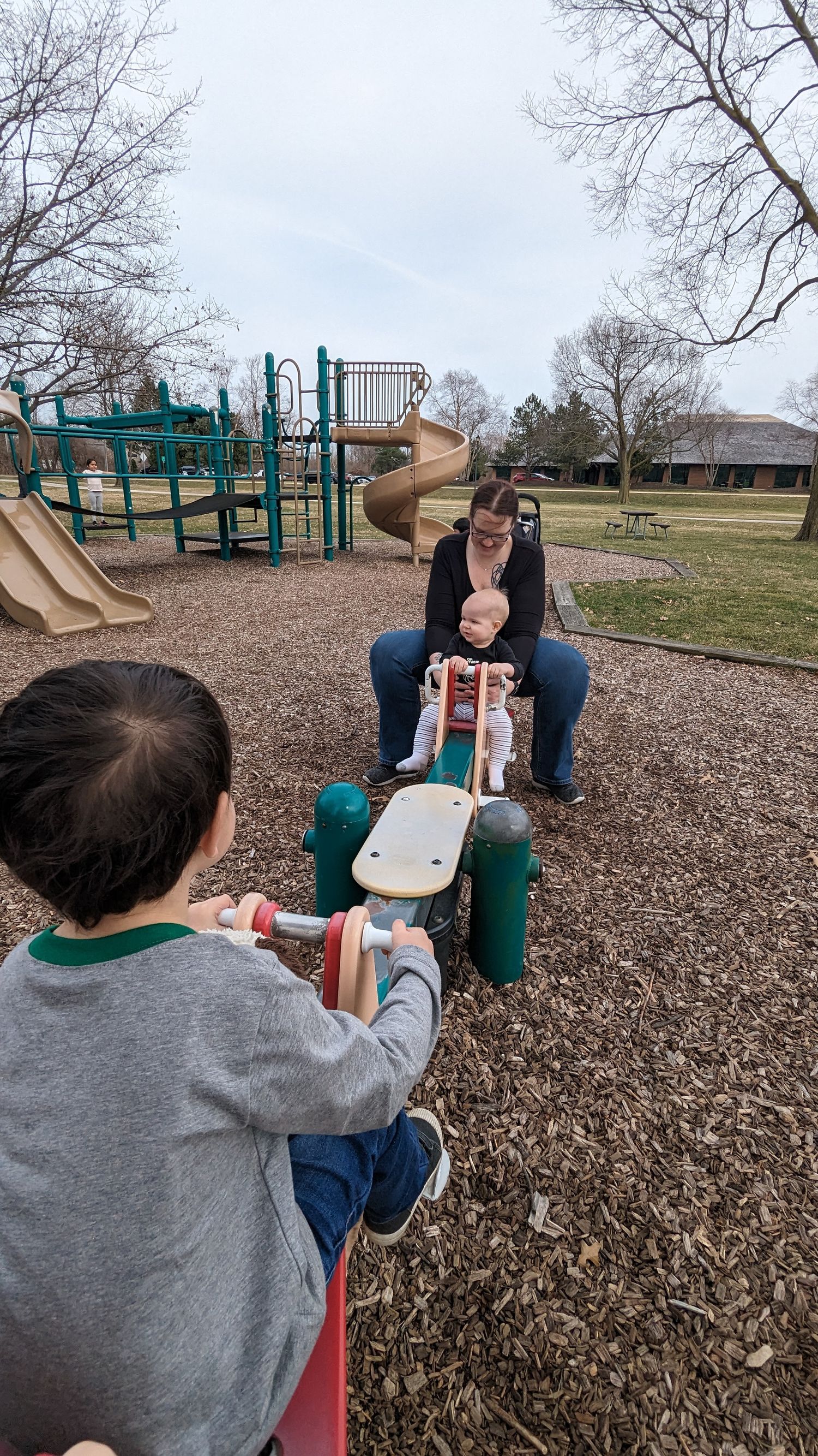 A mother holds her baby on a seesaw while an older child plays at a park playground.