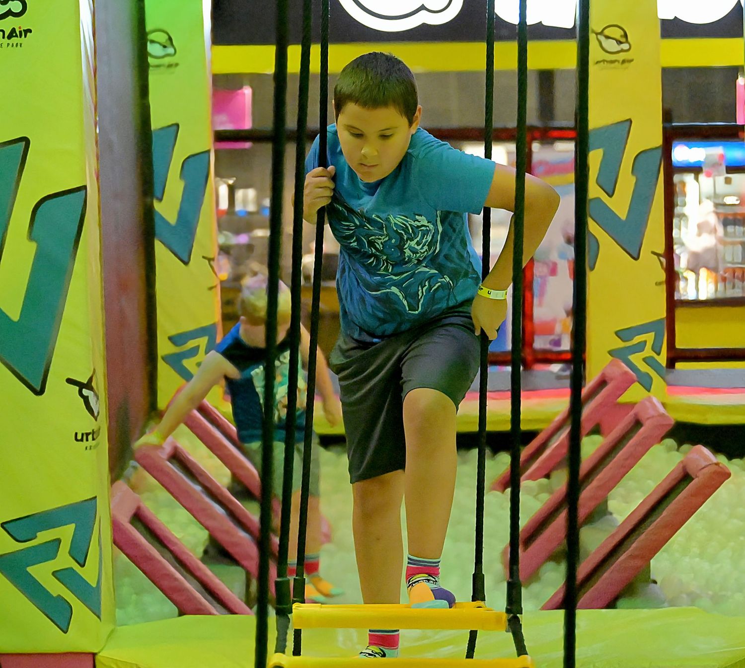 A boy in a blue shirt navigates a rope ladder obstacle course at an indoor adventure park.