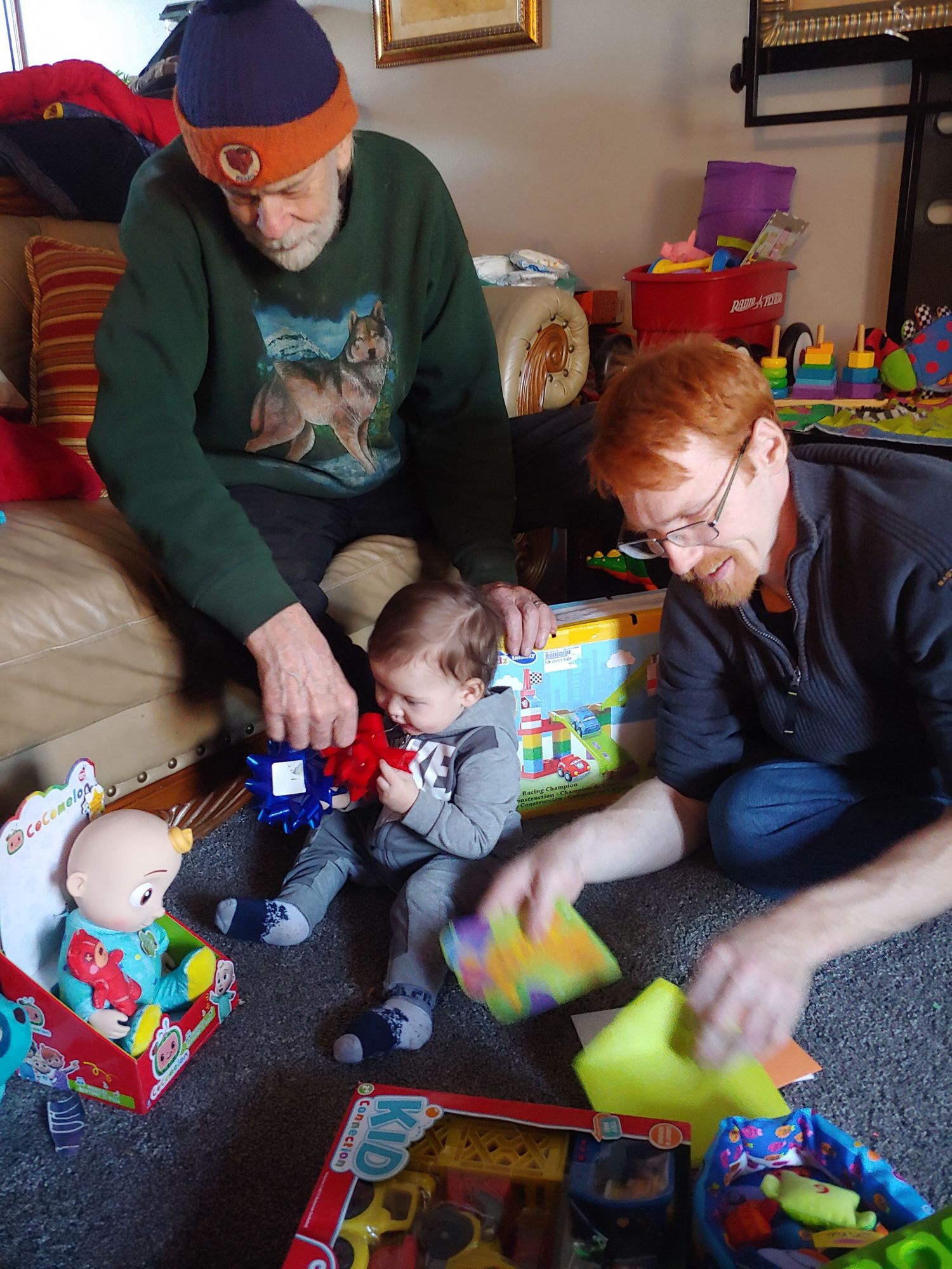 Two men playing with baby surrounded by colorful toys on living room floor.