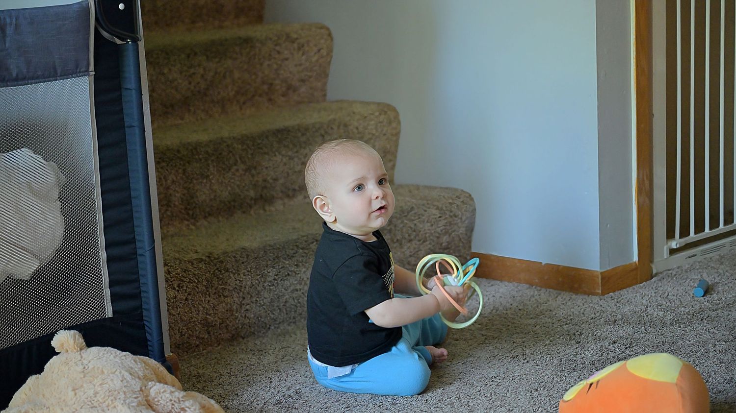 Baby sitting on carpet holding ring toy near staircase with scattered toys.