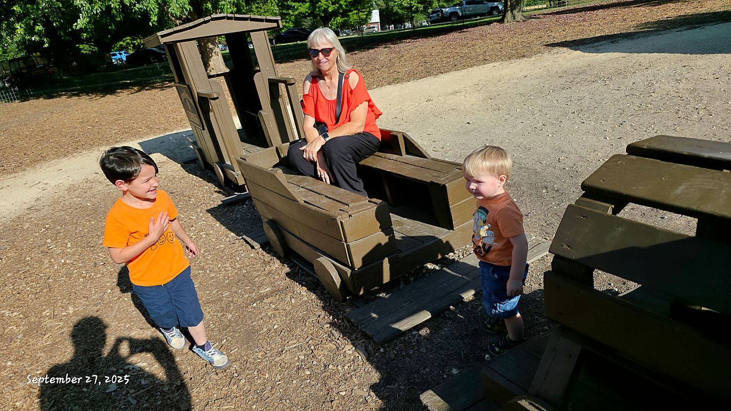 A woman sits on a wooden play train while two young boys play beside her at a park.