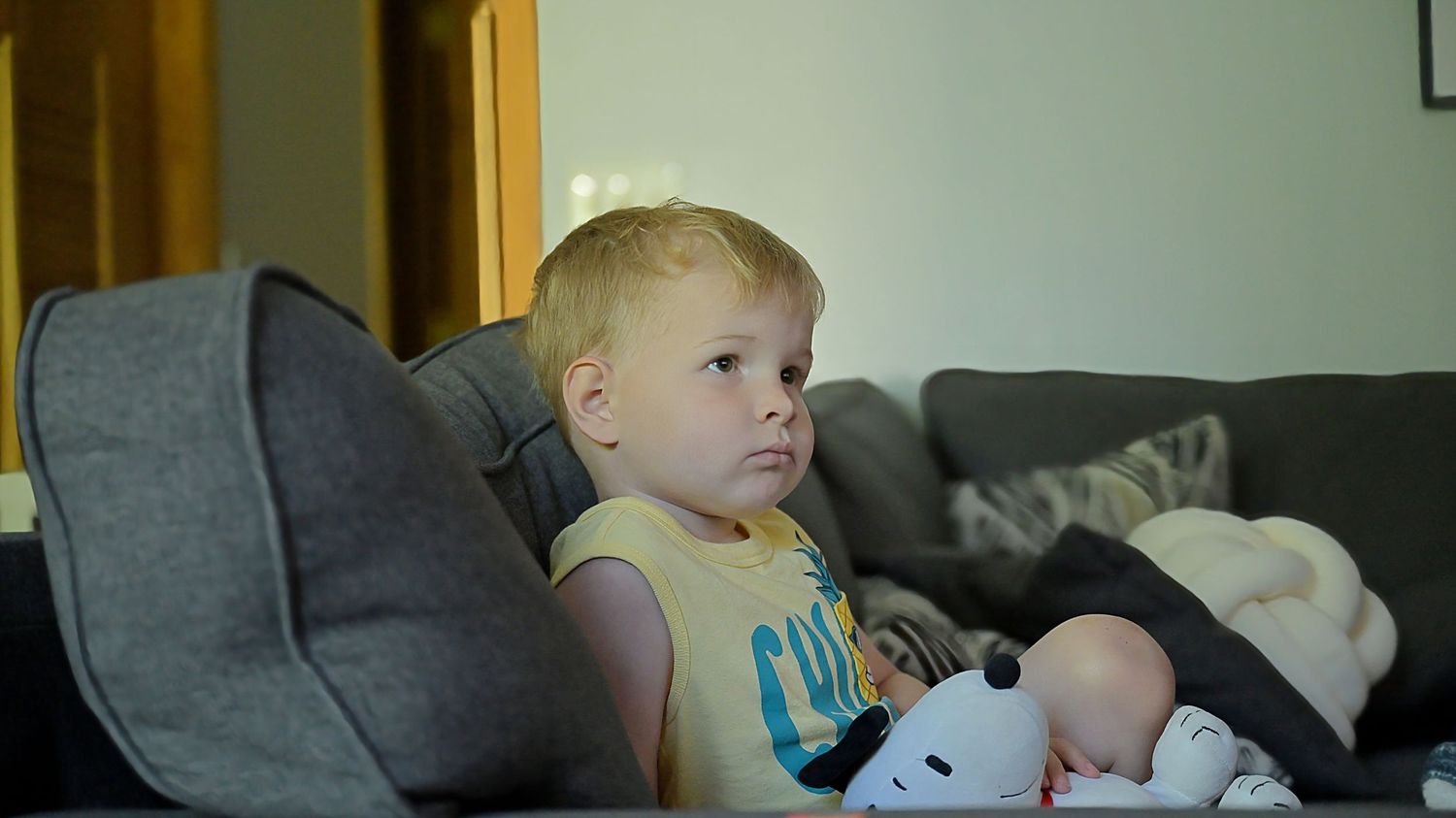 A young toddler sitting on a gray sofa holding a stuffed animal watching television.