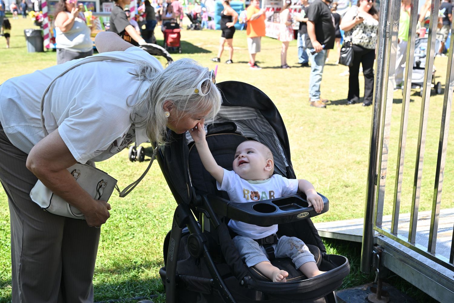 Elderly woman leans down to interact with a laughing baby in a stroller outdoors.