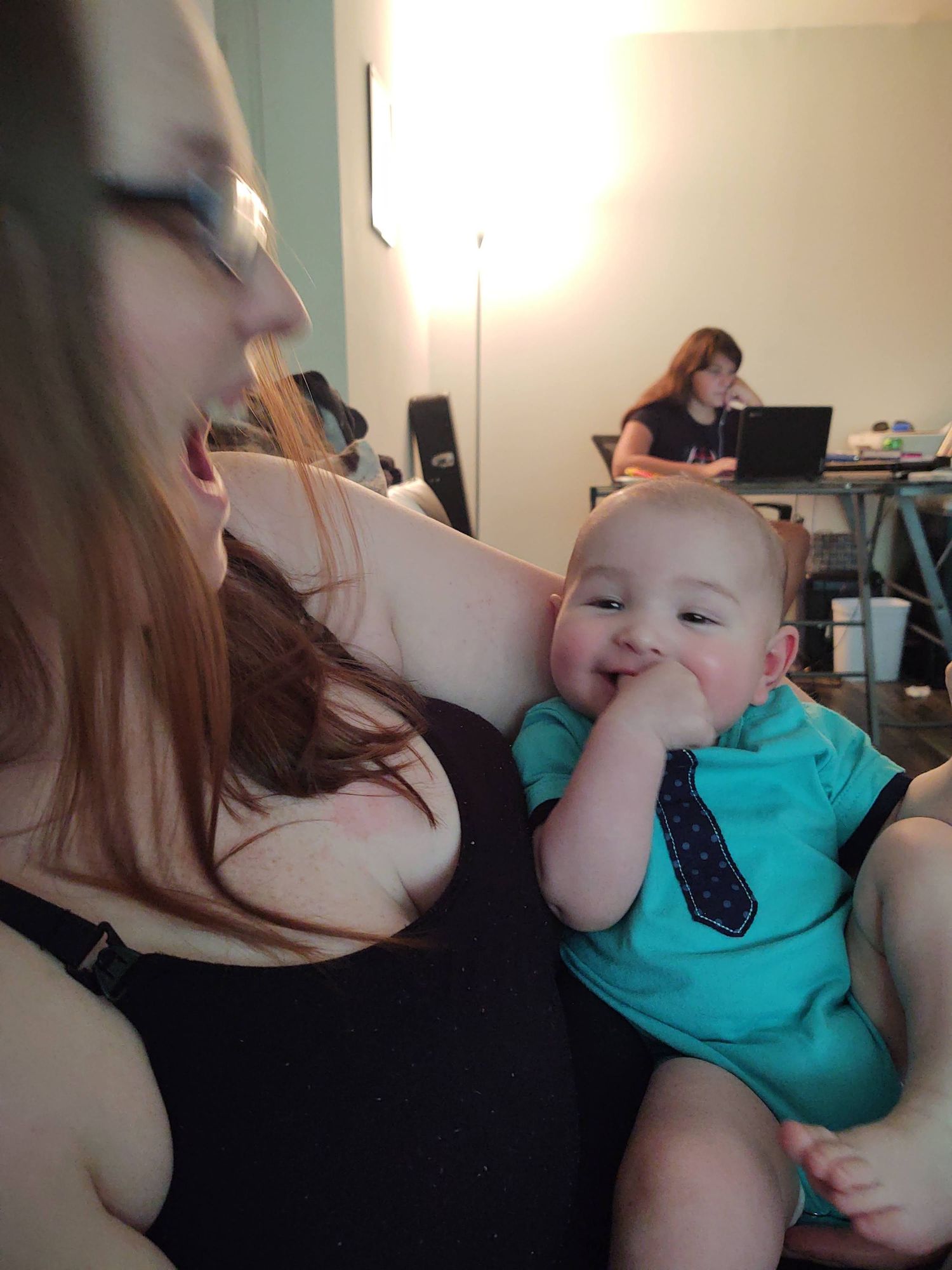 Young mother smiling at happy baby in teal tie onesie sucking fingers.