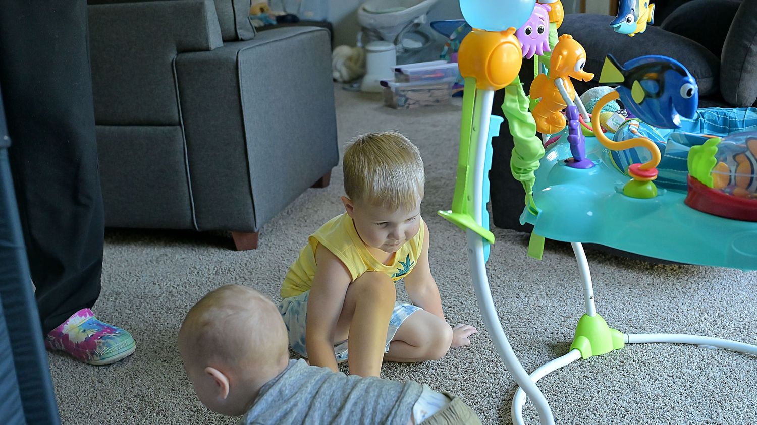 Two young children sit on carpet near a colorful baby activity jumper toy indoors.