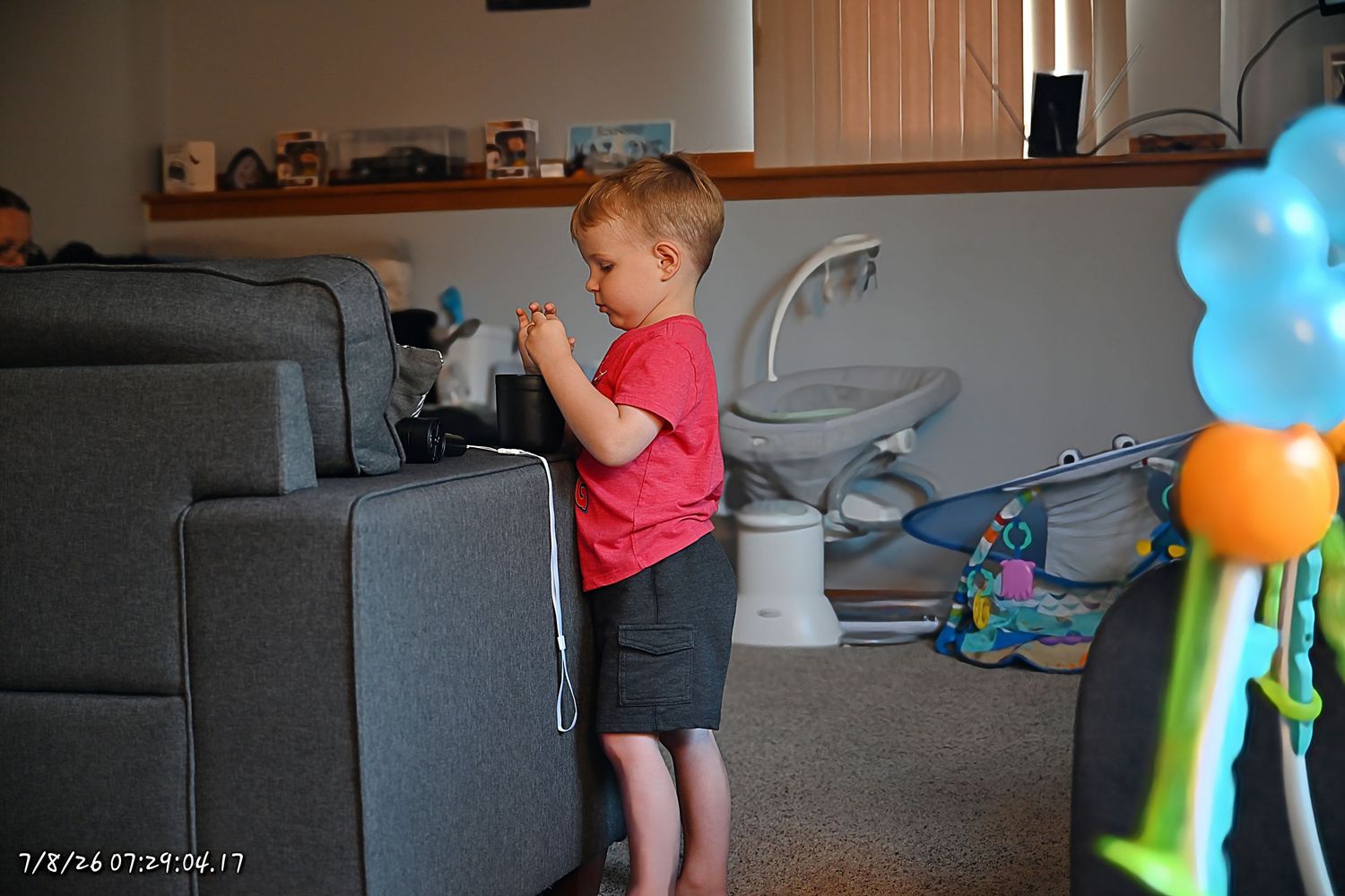 A young boy in a red shirt stands beside a gray sofa in a living room.