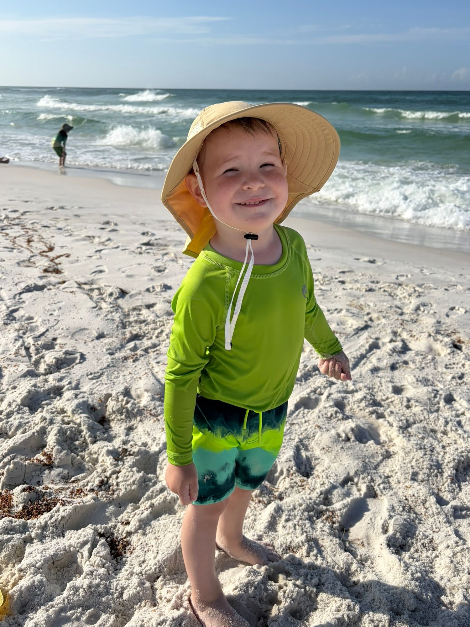 Happy toddler in green rashguard and yellow sun hat smiles on a white sandy beach by the ocean.