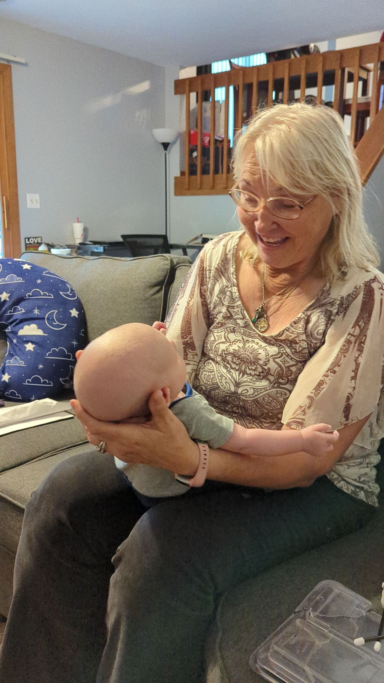 Smiling grandmother with blonde hair and glasses holds newborn baby on couch indoors.