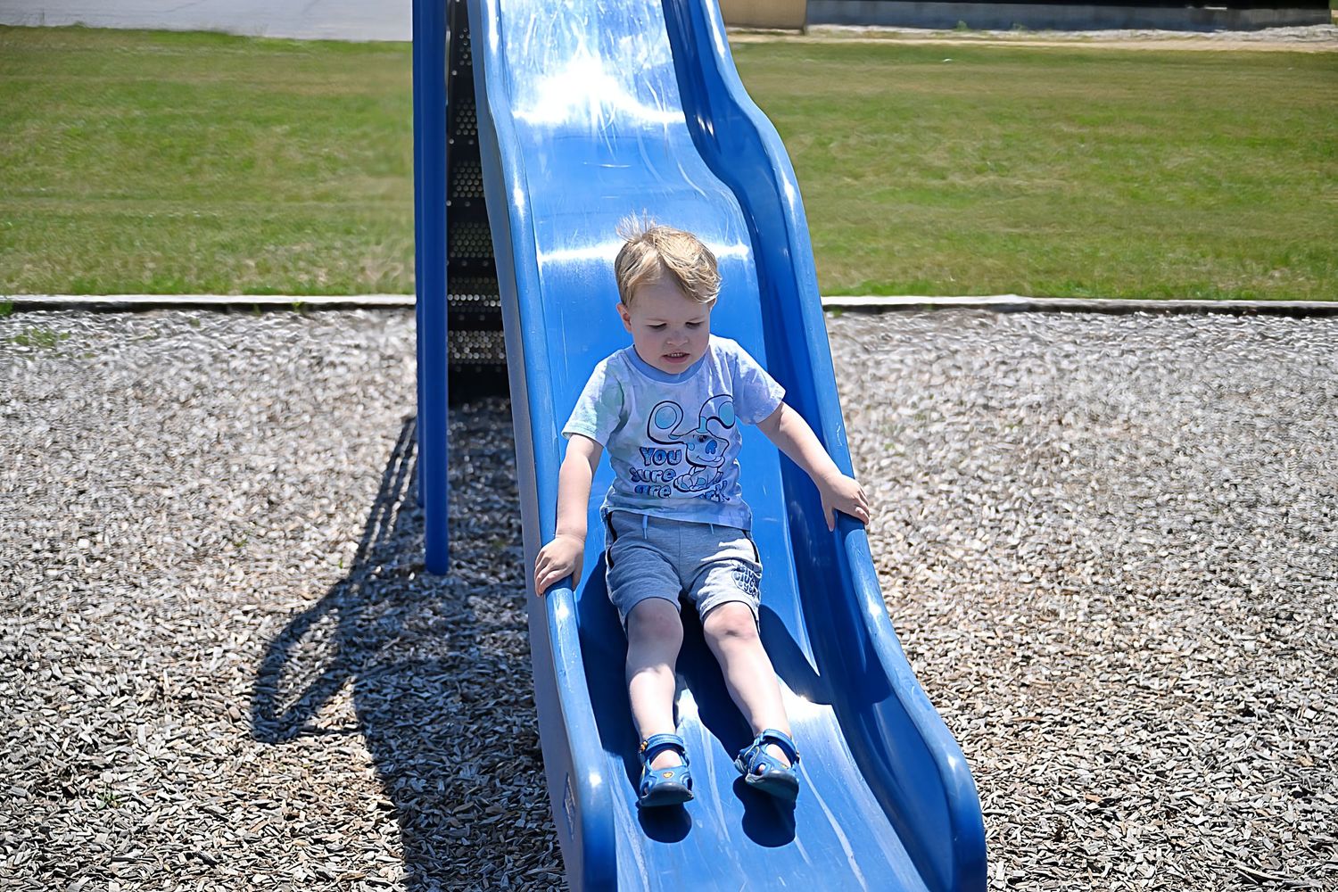A young toddler in a gray shirt slides down a blue plastic slide at an outdoor playground.