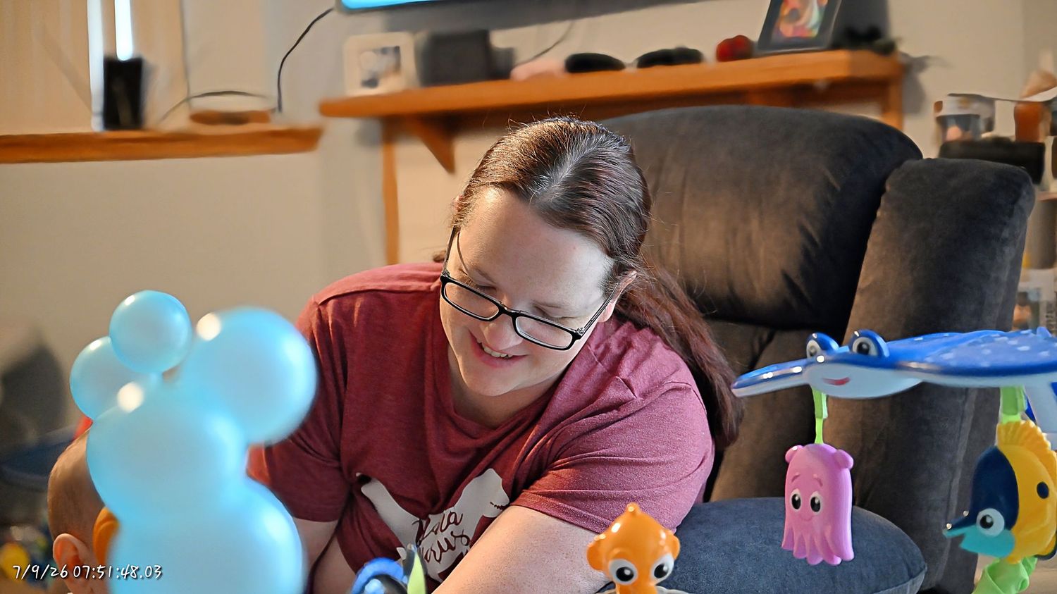 A woman with glasses sitting in a brown recliner playing with colorful baby toys.