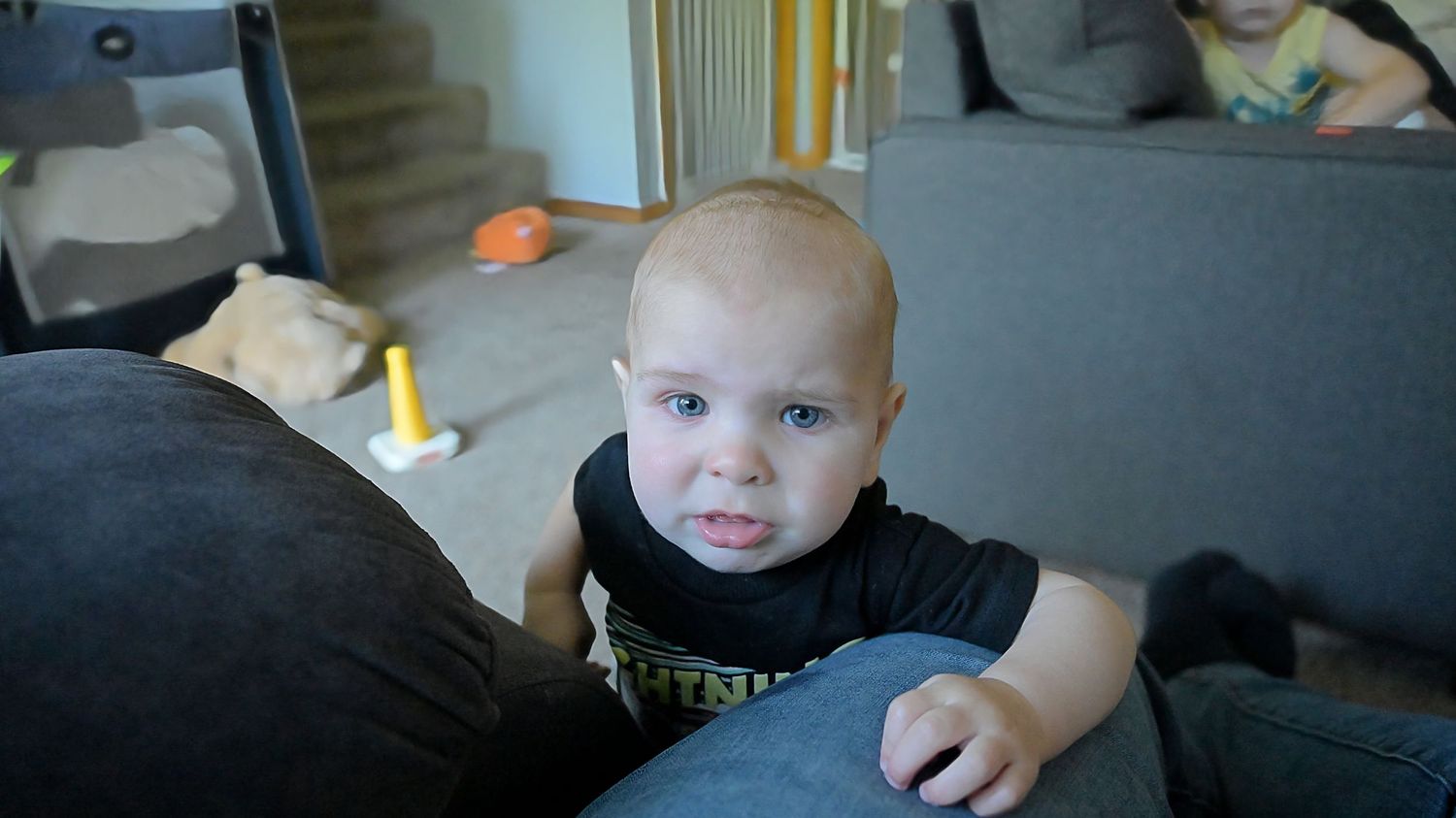 A curious baby in a black shirt looks up while leaning against a gray sofa in a living room.