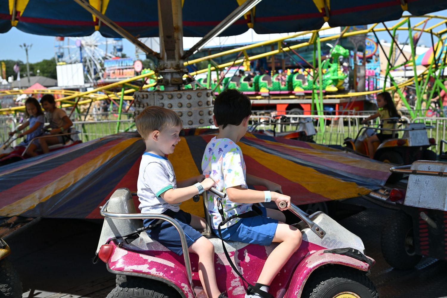 Two boys sharing a carnival car ride at a sunny outdoor amusement fair.