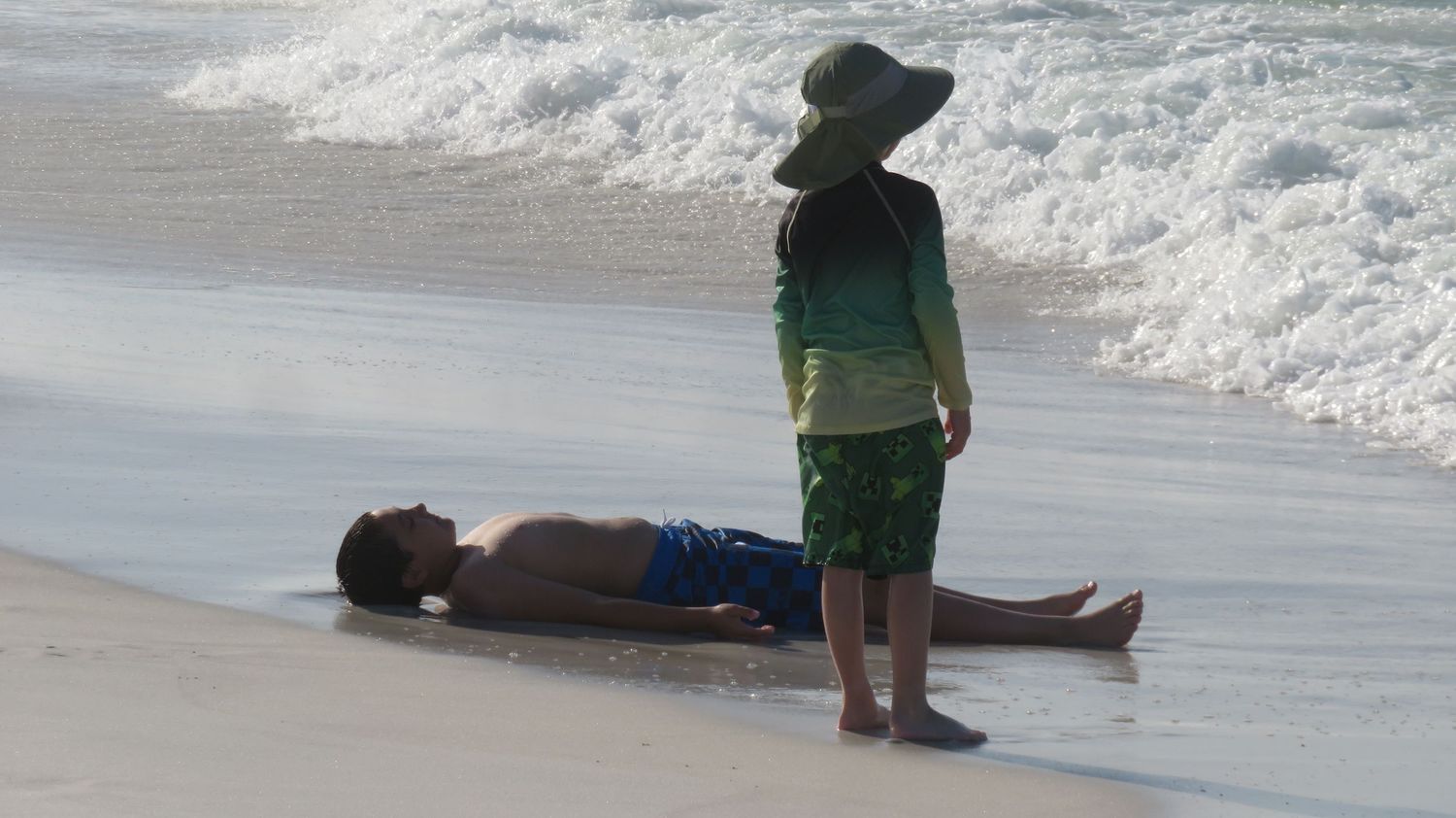 A child in green rash guard stands near a man lying on the wet sandy beach shoreline.