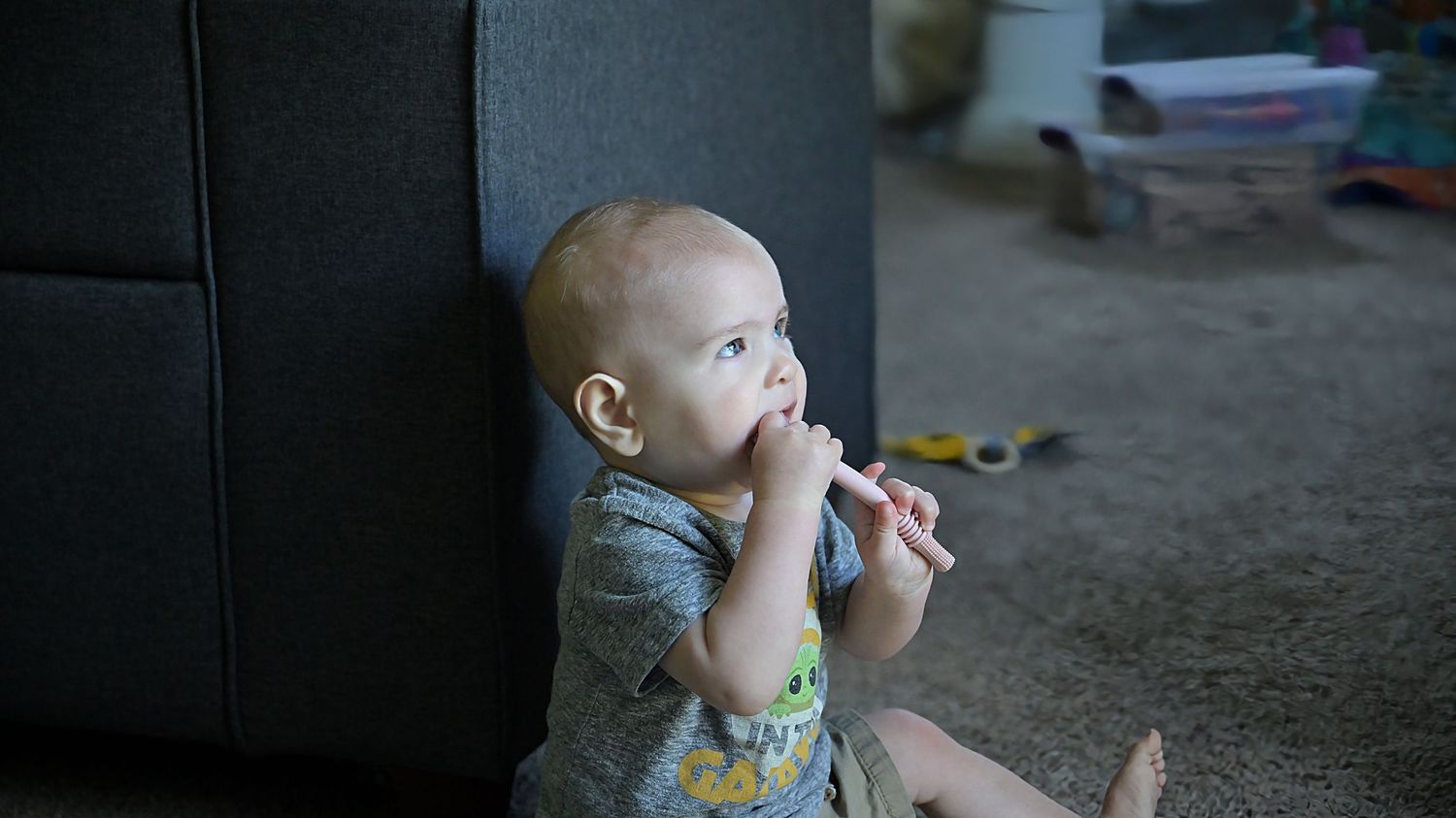 A baby sits against a gray sofa on carpet, chewing on a small object while looking up.