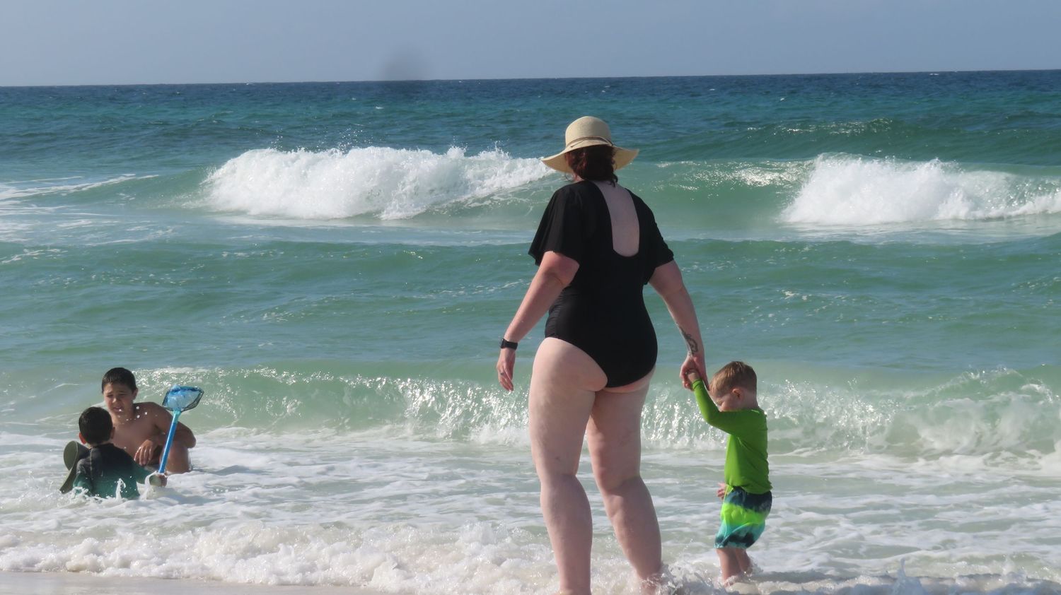 Woman in black swimsuit holds toddler's hand while another child plays in ocean waves.