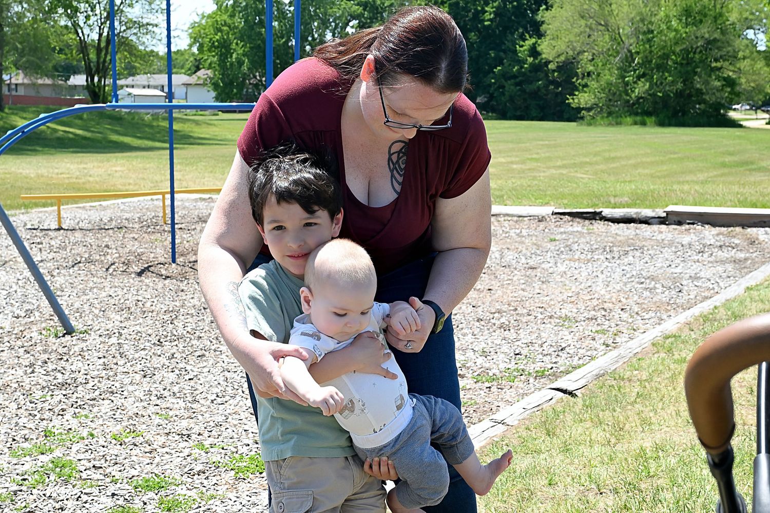 Mother helping young boy hold a baby on a playground swing on a sunny day.