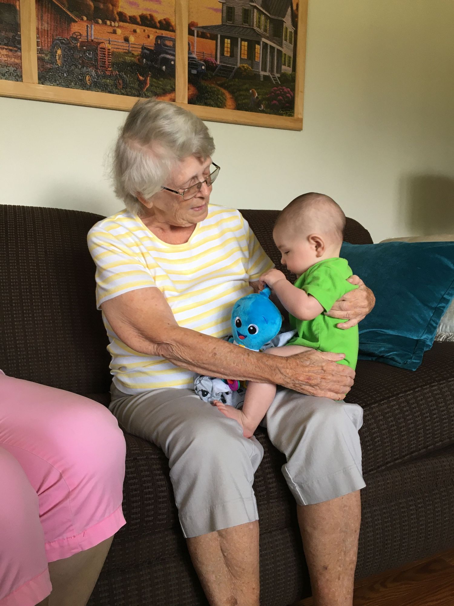 Elderly grandmother sitting on couch holding infant in green outfit with blue toy.