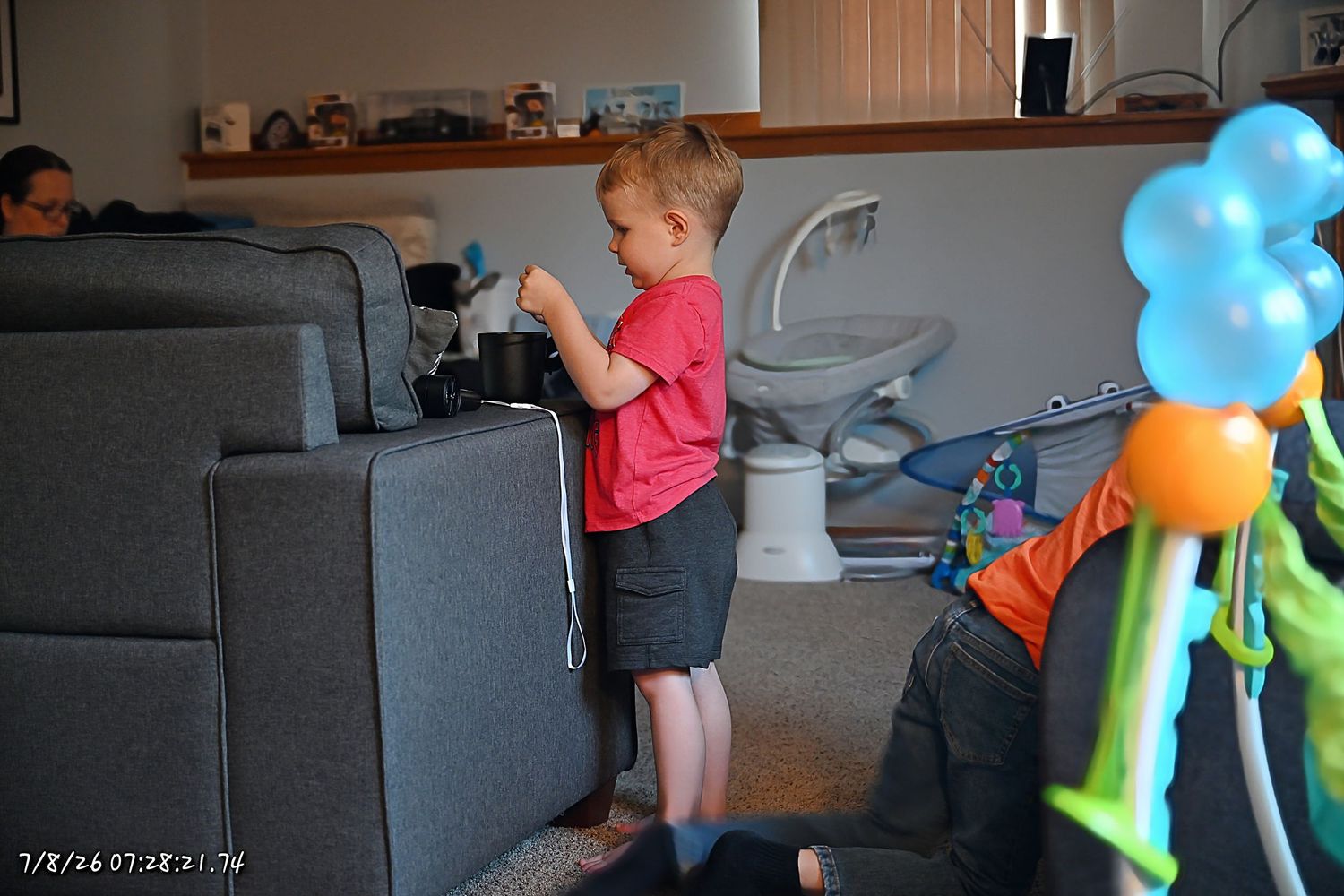 A young boy in a red shirt stands by a gray sofa with colorful balloon decorations nearby.