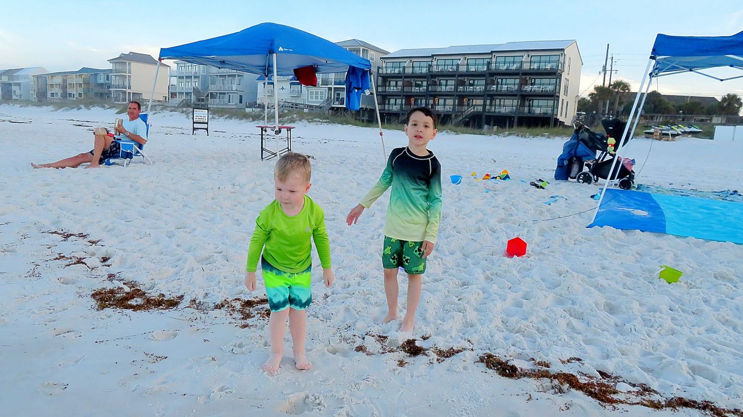 Two young boys in colorful rashguards play on a white sandy beach with blue tents and ocean resort backdrop.