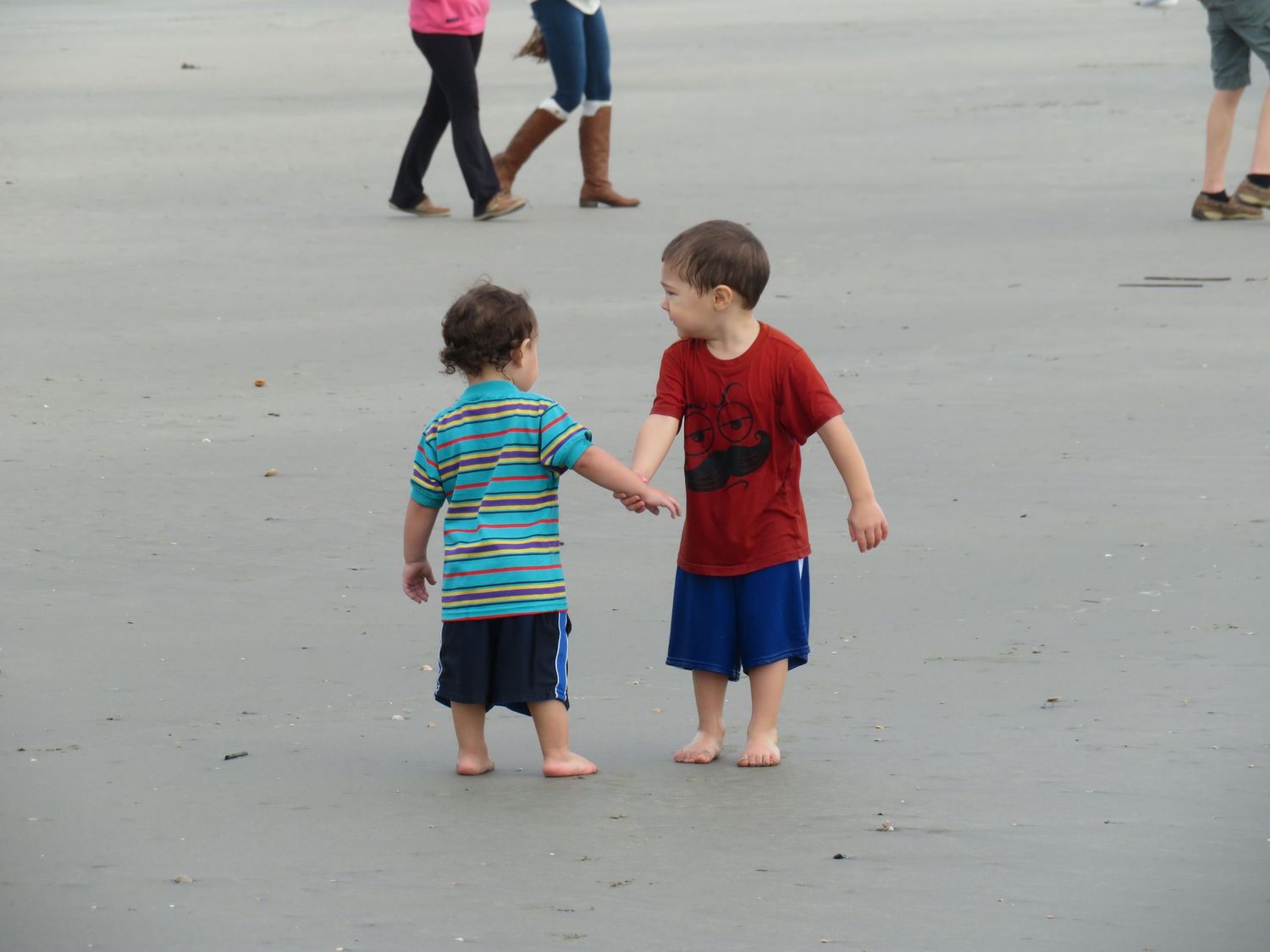 Two young boys holding hands on a sandy beach shore.