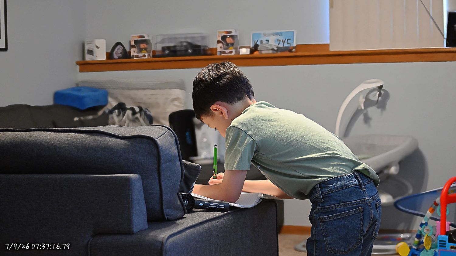 A young boy in a green shirt leaning over a blue couch looking at something in a room.