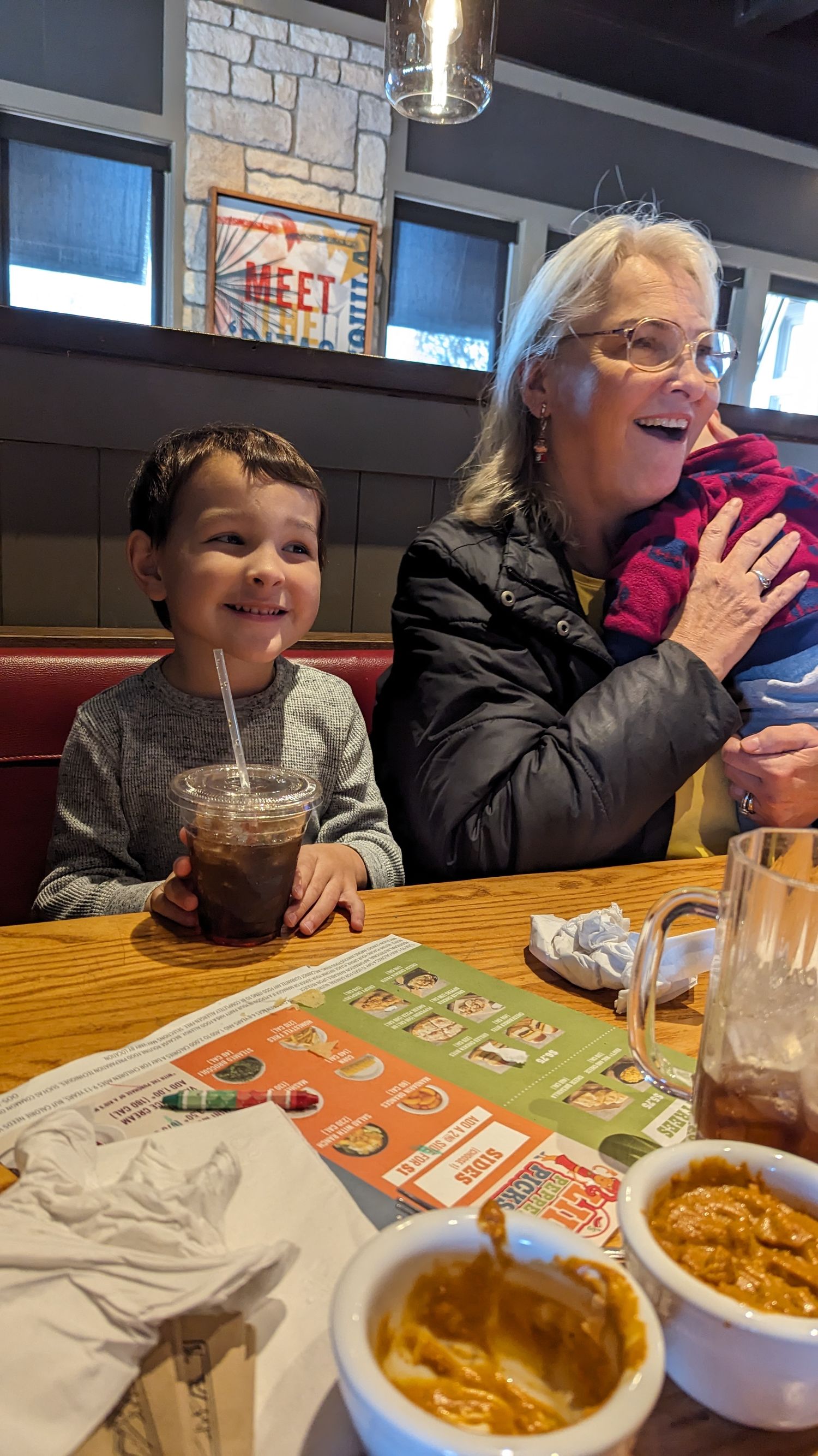 Smiling young boy with iced drink sits beside animated grandmother at restaurant booth.