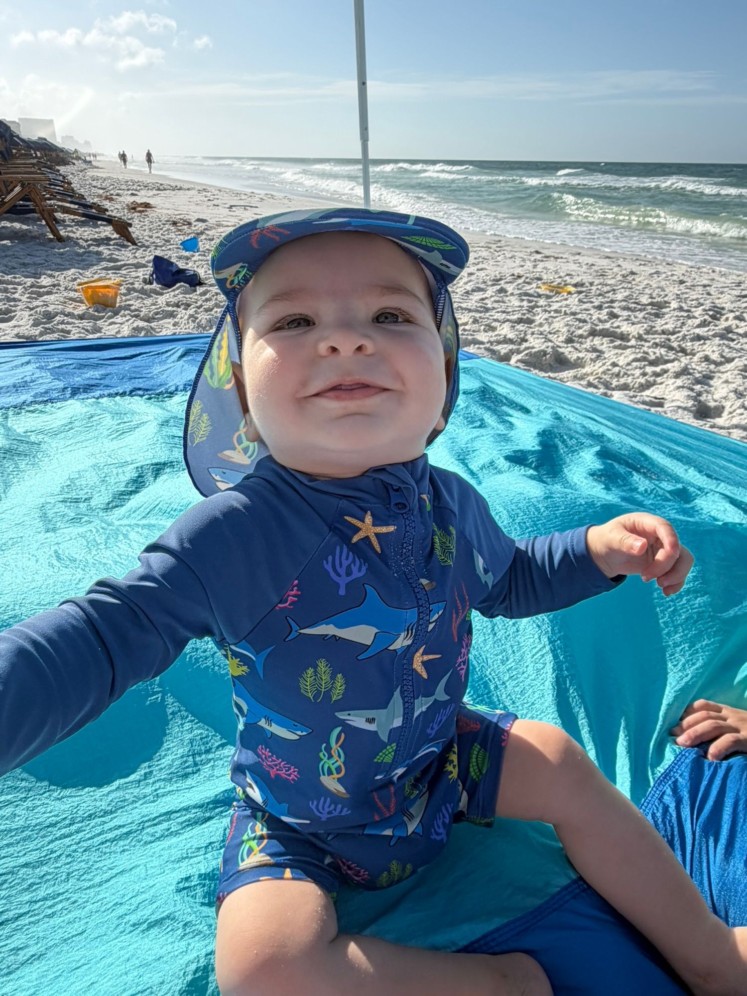 Adorable baby in blue marine life swimsuit and sun hat sits on beach blanket by the ocean.