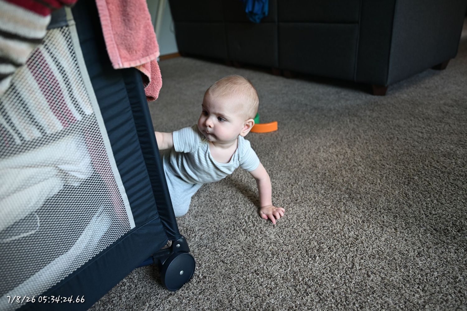 A curious baby in a light blue onesie pulls up to stand against a mesh playpen on carpet.
