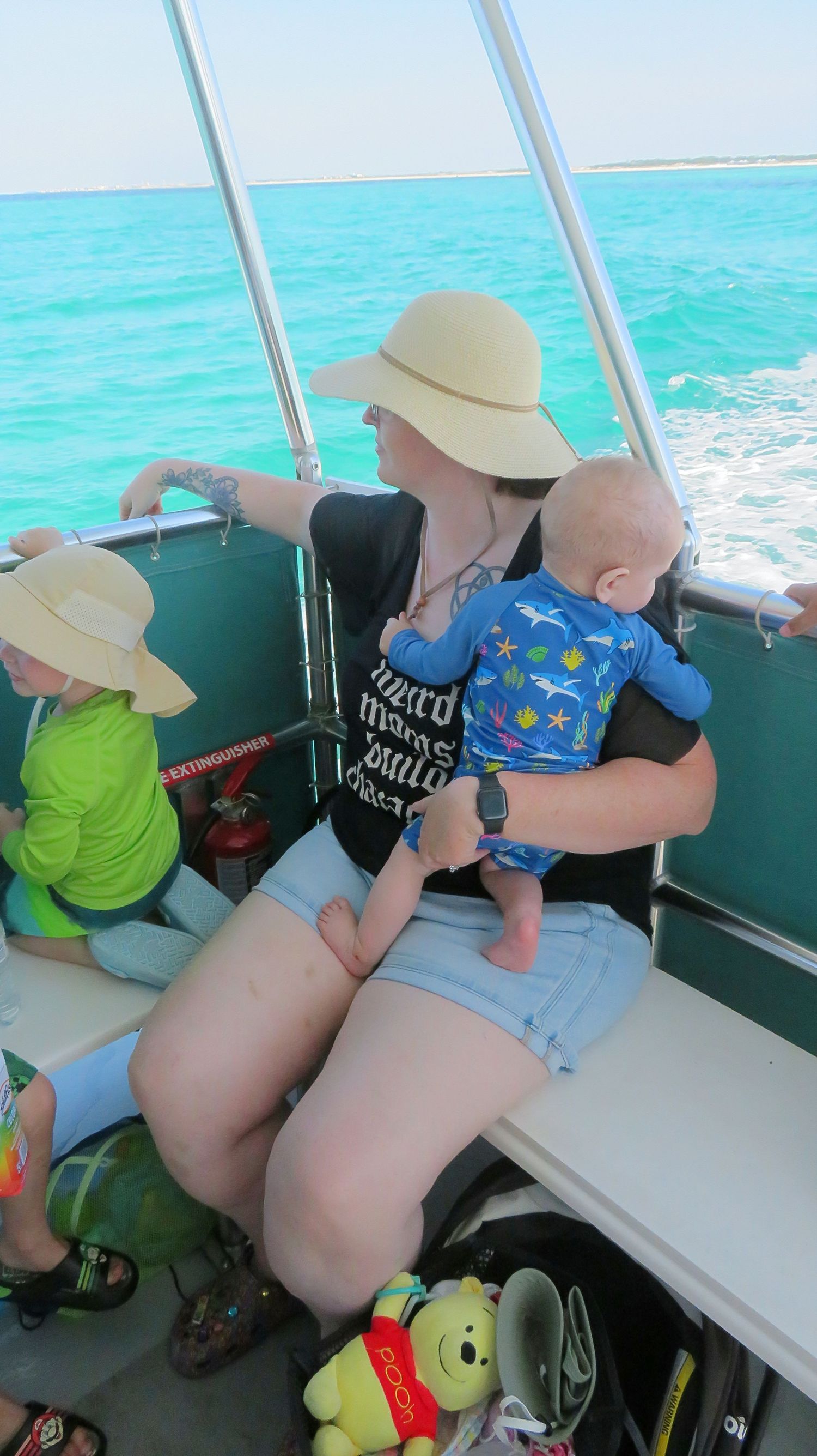 Mother in sun hat holding baby in blue outfit on a boat with turquoise ocean views.