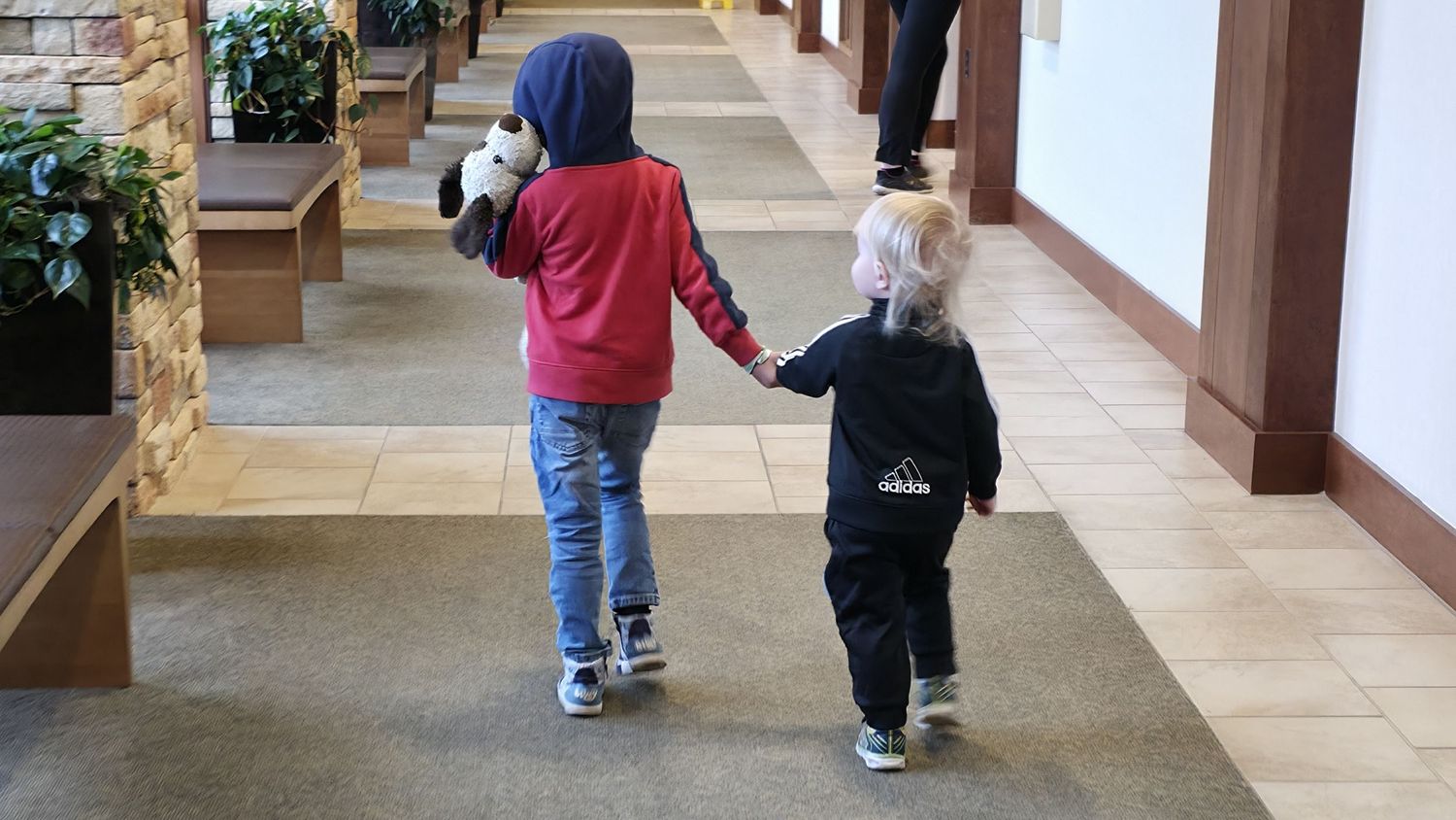 Two young children walk hand in hand down a warmly lit indoor hallway.
