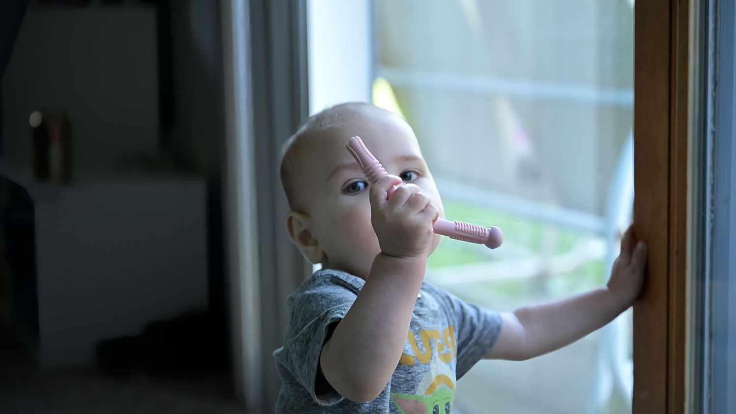 A curious baby stands at a window, holding a small object to their mouth while looking outside.