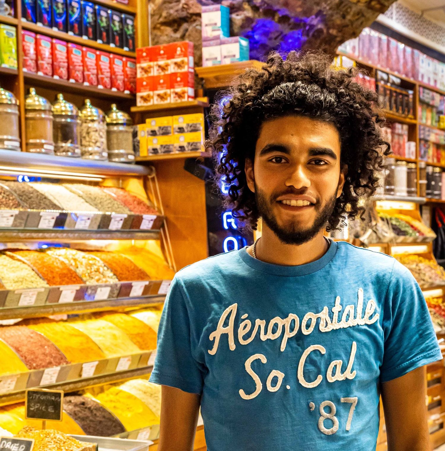 Young man in Aeropostale t-shirt smiling in colorful spice market shop.