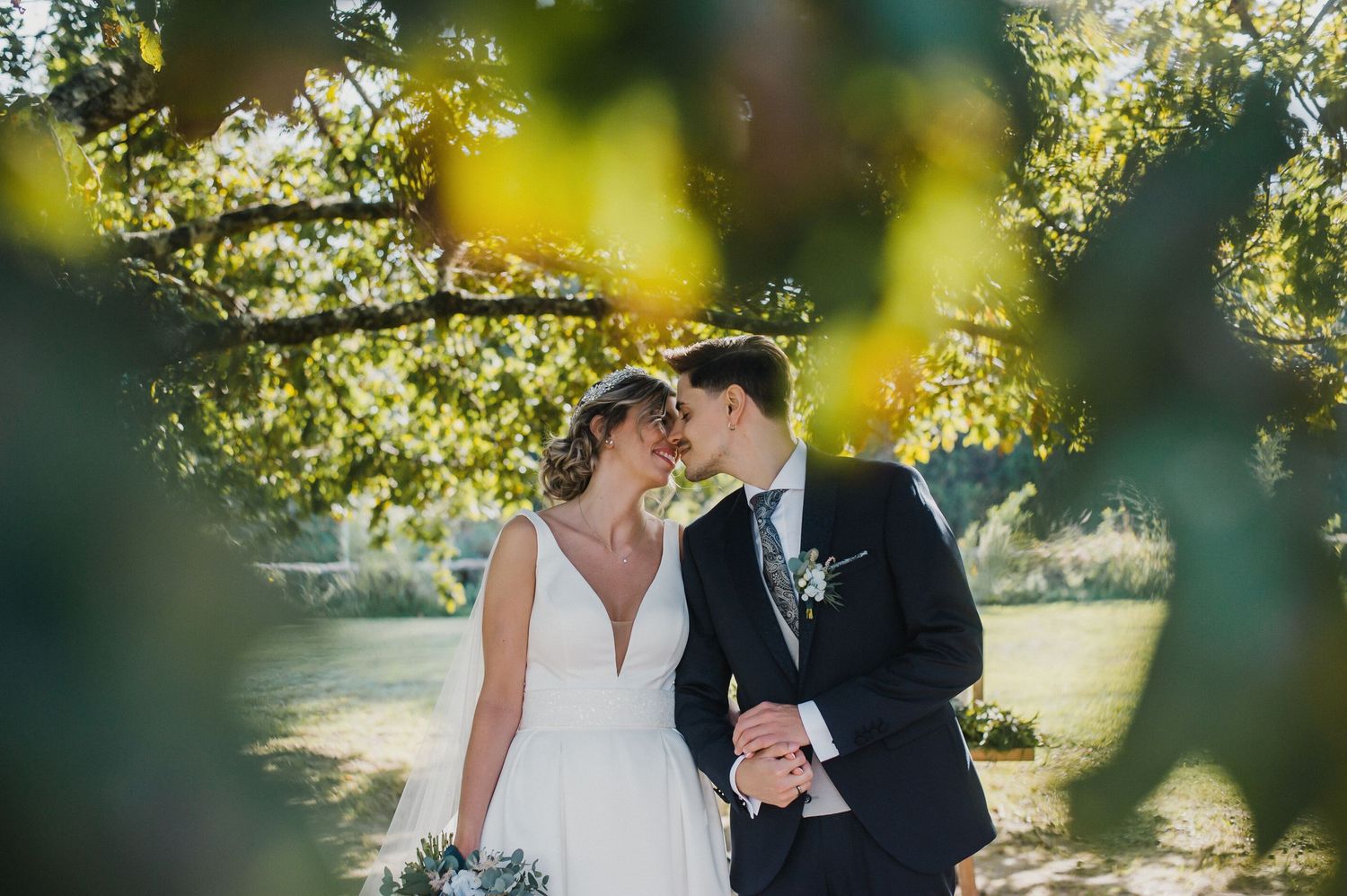 Newlywed couple sharing an intimate moment under a sunlit tree in a lush garden setting.