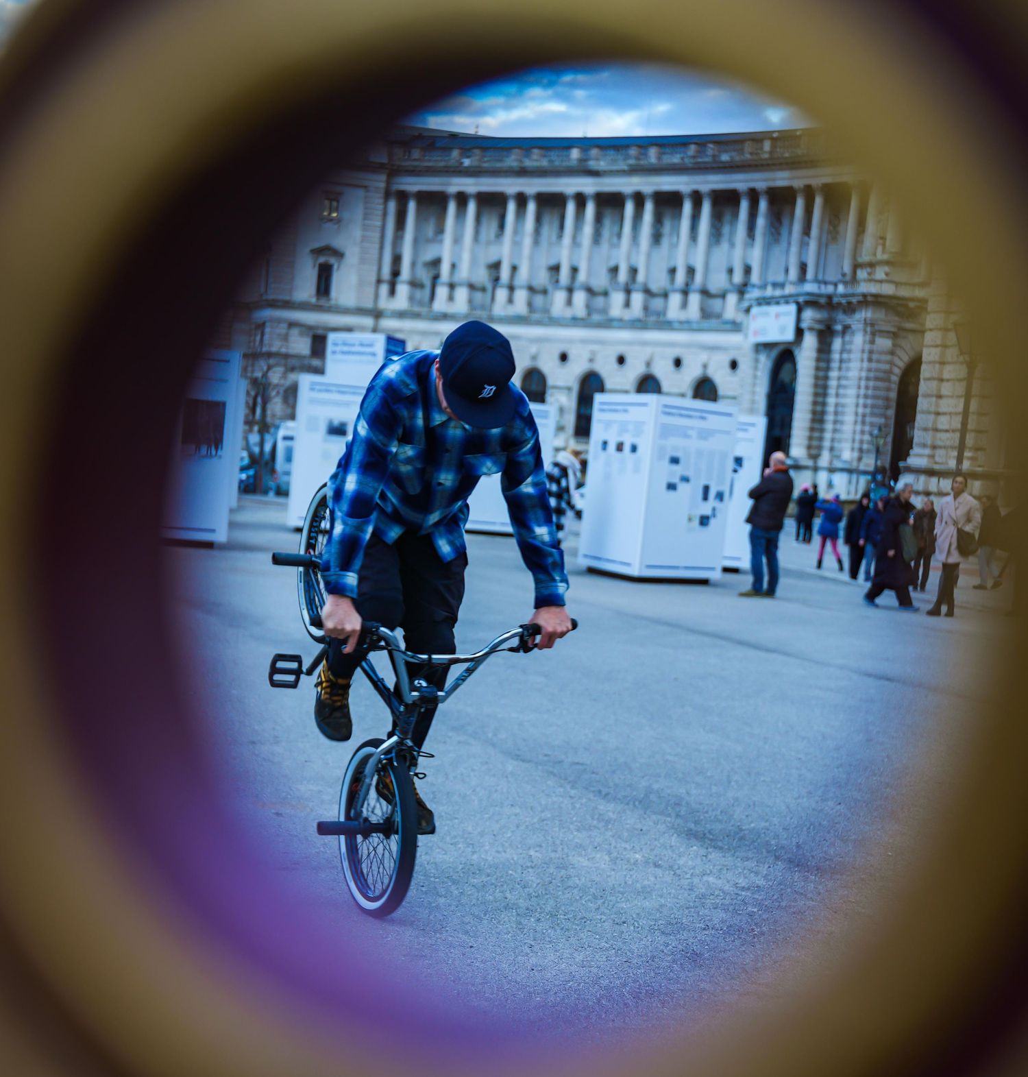 BMX rider performing tricks in a city square viewed through a circular frame or tube.