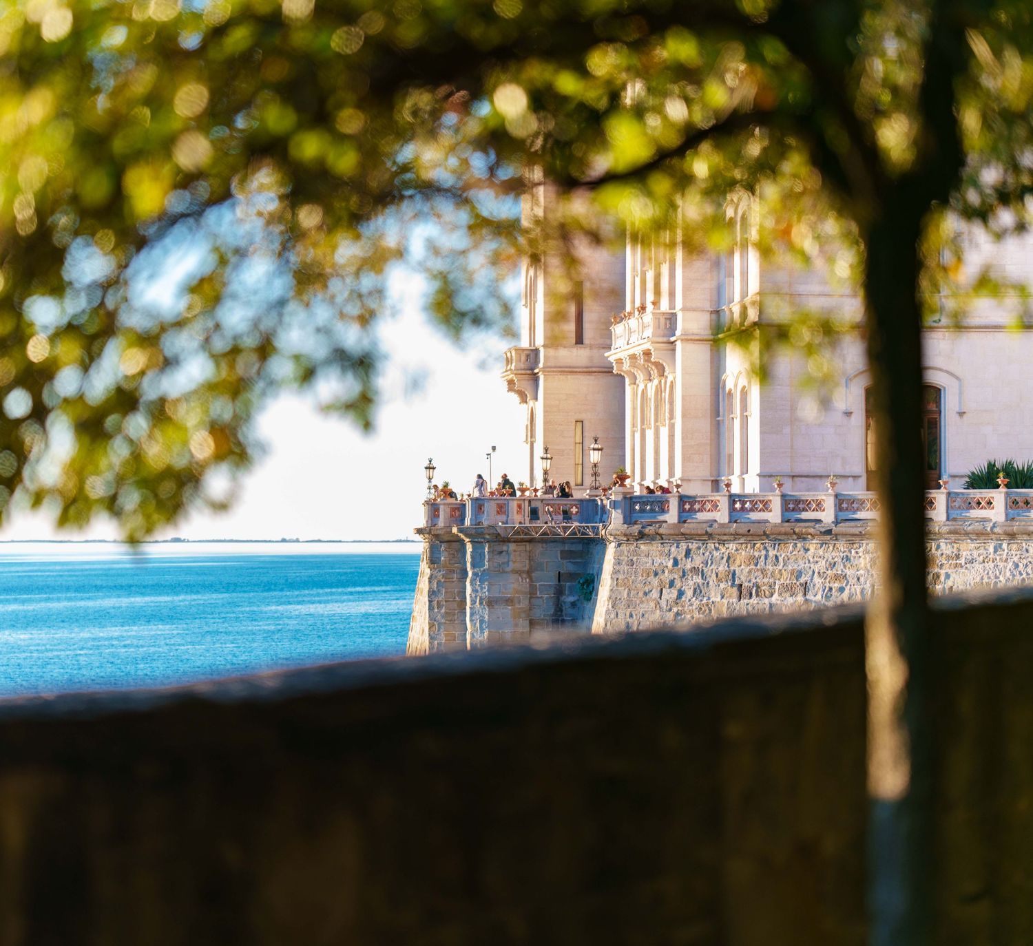 Scenic view of a historic waterfront palace framed by tree branches with calm blue water in background.