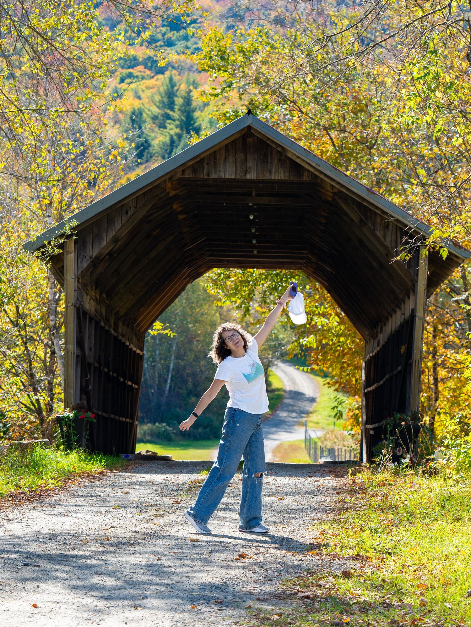 Joyful woman posing with arms outstretched at a rustic covered bridge surrounded by autumn foliage.