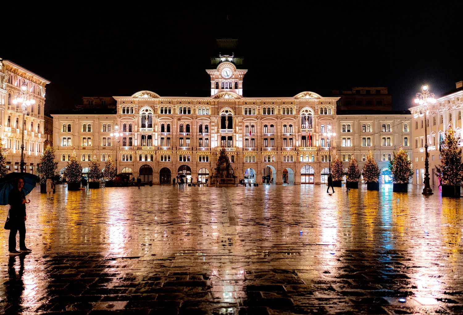 A grand illuminated government building reflects on wet cobblestones in Triestes Piazza Unita at night.
