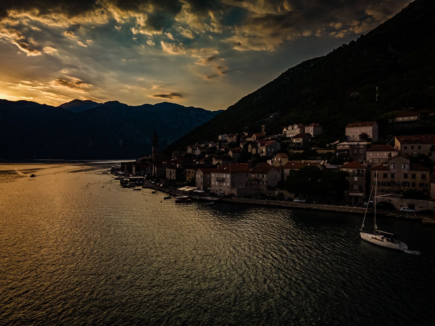 A dramatic sunset illuminates a historic coastal village nestled between mountains and calm waters in Montenegro.
