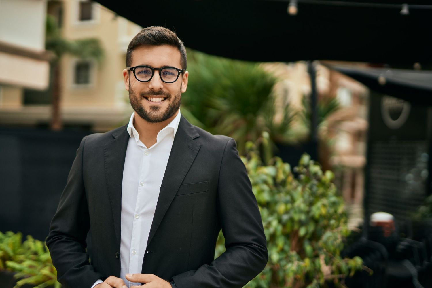 Smiling young man in glasses and black suit jacket outdoors near palm trees.